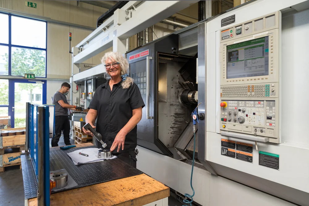 Mit einem digitalen Dreipunkt-Innenmessgerät kontrolliert Zerspanungsmechanikerin Birgit Pawellek, ob die vorgegebenen Maße bei den im Dreikanal-Universal Dreh- und Fräs-Zentrum bearbeiteten Werkstücken eingehalten worden sind. Im Hintergrund bereitet Zerspanungsmechaniker und Ausbilder David Bellavona weitere Werkstücke vor, die anschließend in dem CNC-Bearbeitungszentrum nach individuellen Kundenwünschen bearbeitet werden.
