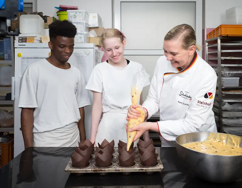 Karin Steinhoff zeigt Anna Brinkmann und Amadou Bah, wie sie die sogenannten Tulpen mit Muffinteig füllt. Traumberufe Bäcker und Konditor 2026