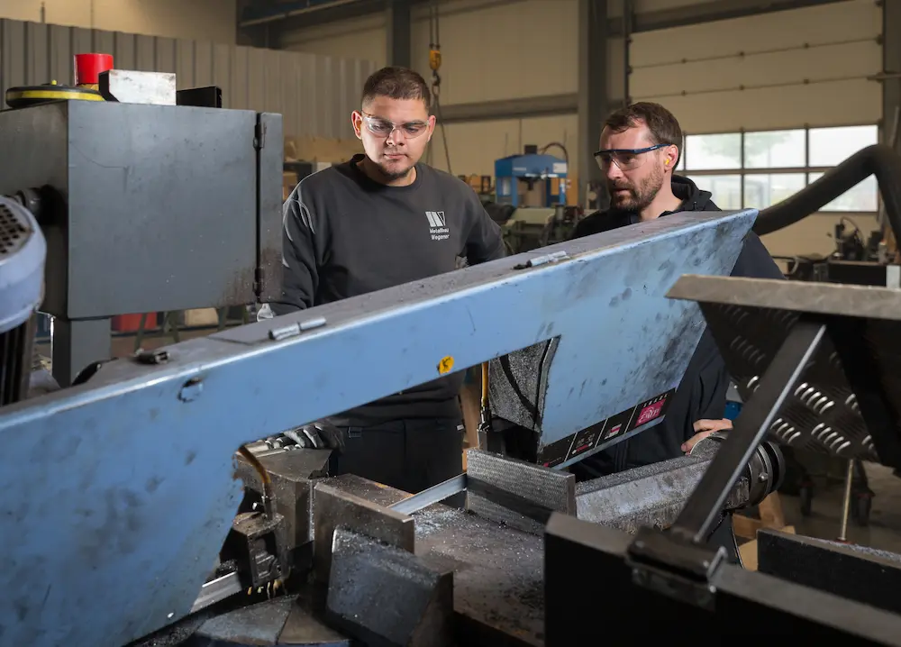 Mit einer Bandsäge sägt der angehende Metallbauer Anil Deveci ein Flachstahlelement auf die geforderte Länge, während ihm Metallbauermeister Jonas Milewzyk dabei über die Schulter schaut und Tipps gibt.