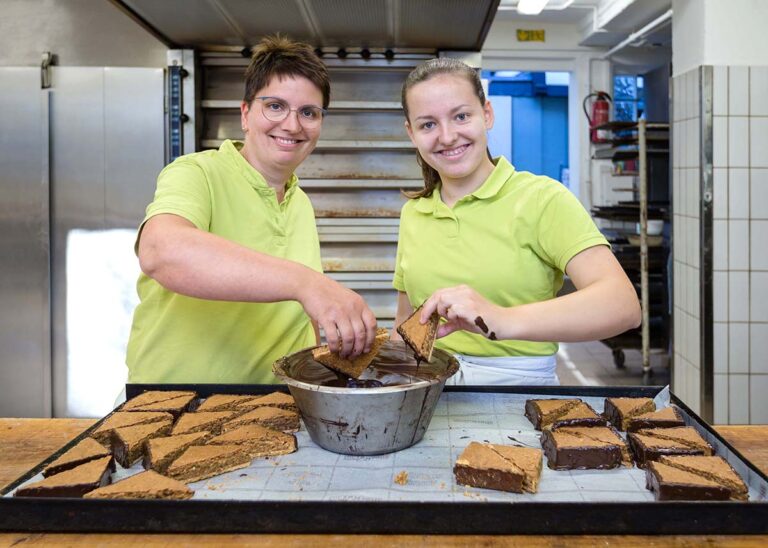Konditor- und Bäckermeisterin Vera Christiani taucht gemeinsam mit der Auszubildenden Paula Dahlhoff Nussecken in flüssige Schokolade und gibt ihr dabei Tipps. (Foto: Gabi Bender)