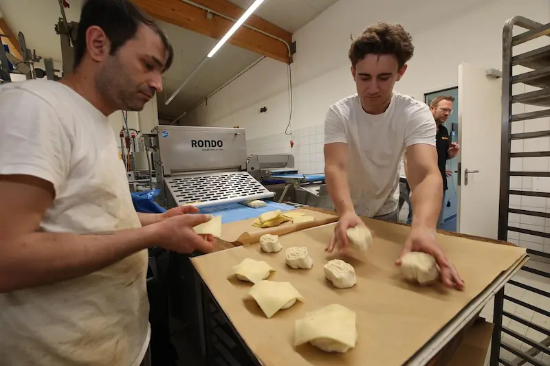 Die Auszubildenden Tim Braune (rechts) und Maslun Aksan belegen Blätterteigrollen mit Käse zum Überbacken. Traumberufe Bäcker und Konditor 2026