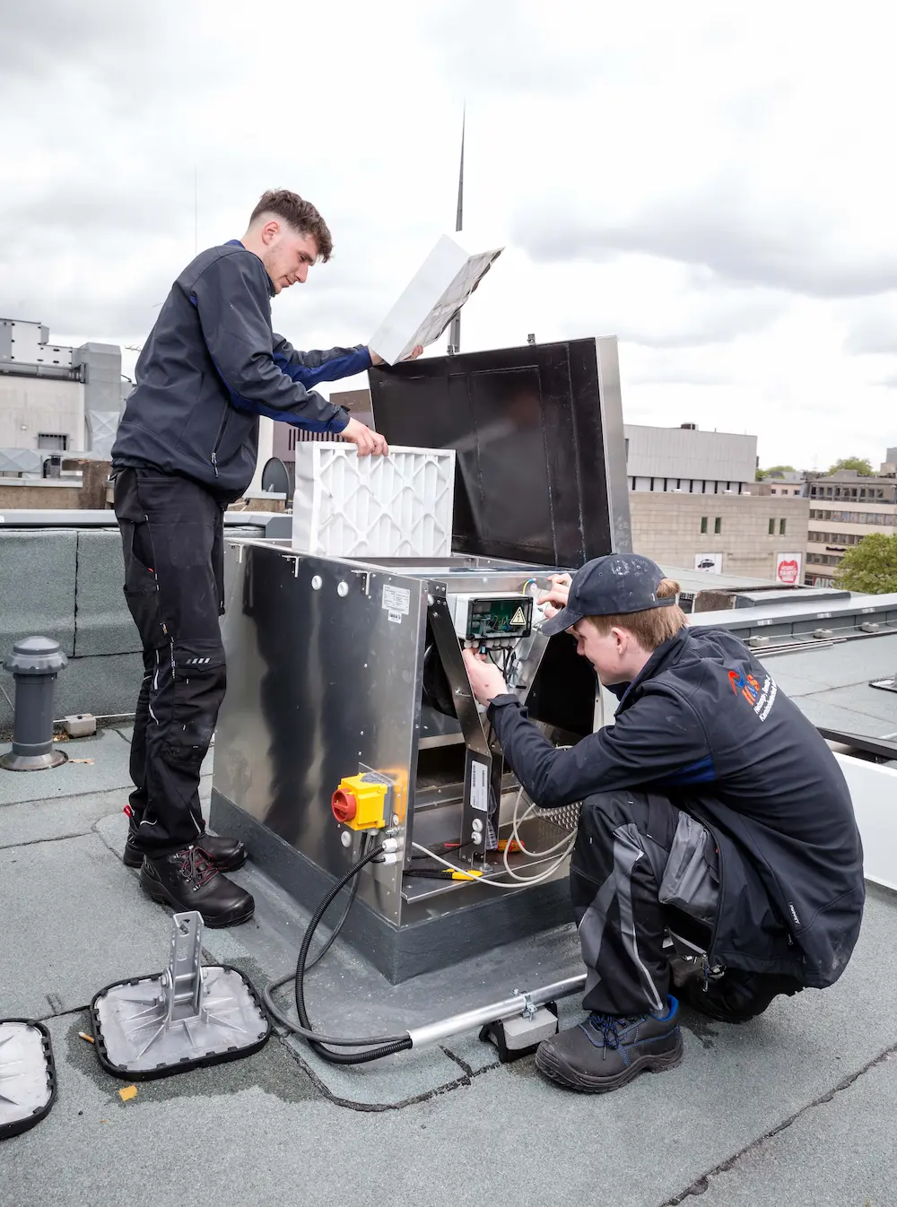 Über den Dächern von Dortmund setzt Anlagenmechaniker Till Sören Bönke (links) einen neuen Luftfilter bei einer Lüftungsanlage ein, während Azubi Piet Bremen vorsorglich die Kabelverbindungen überprüft. Traumberufe 2026 Anlagenmechaniker für SHK